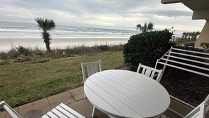 Un patio con una mesa redonda y sillas blancas, con vista a una playa y olas del océano, además de palmeras.