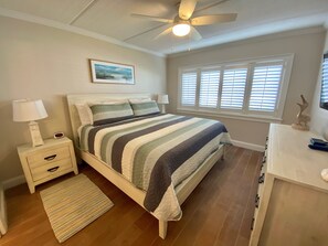 Une chambre à coucher comprenant un lit, des tables de chevet, une commode, un ventilateur de plafond, une fenêtre avec des stores et une œuvre d’art accrochée au mur.
