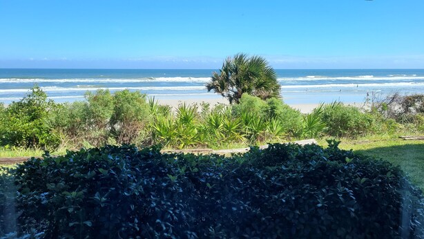 Vista del océano desde una ventana con vistas a una playa y vegetación densa al frente.