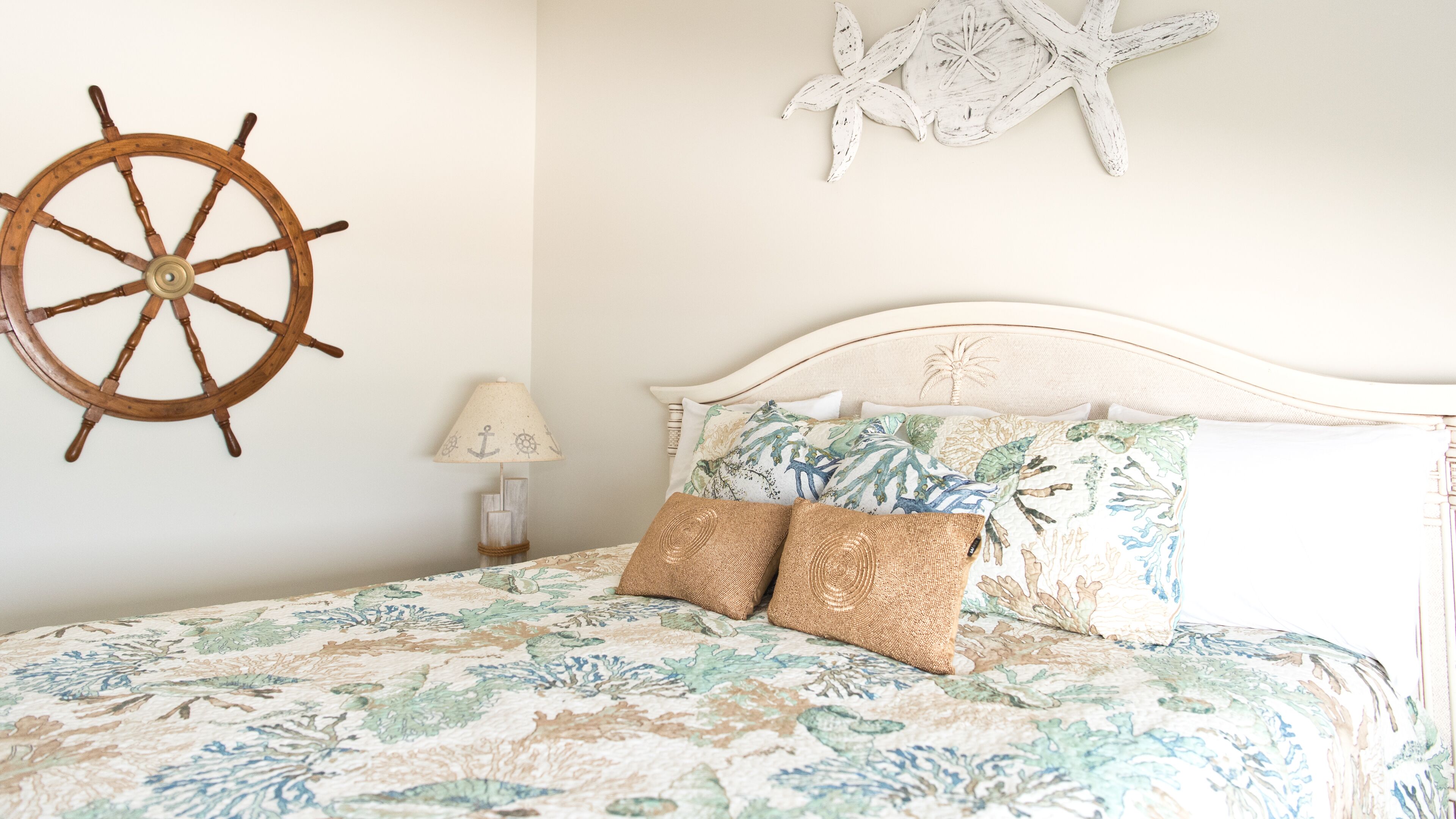 A bed with a floral bedspread, two pillows, a wooden ship wheel, a starfish decoration, and a lamp on a nightstand.