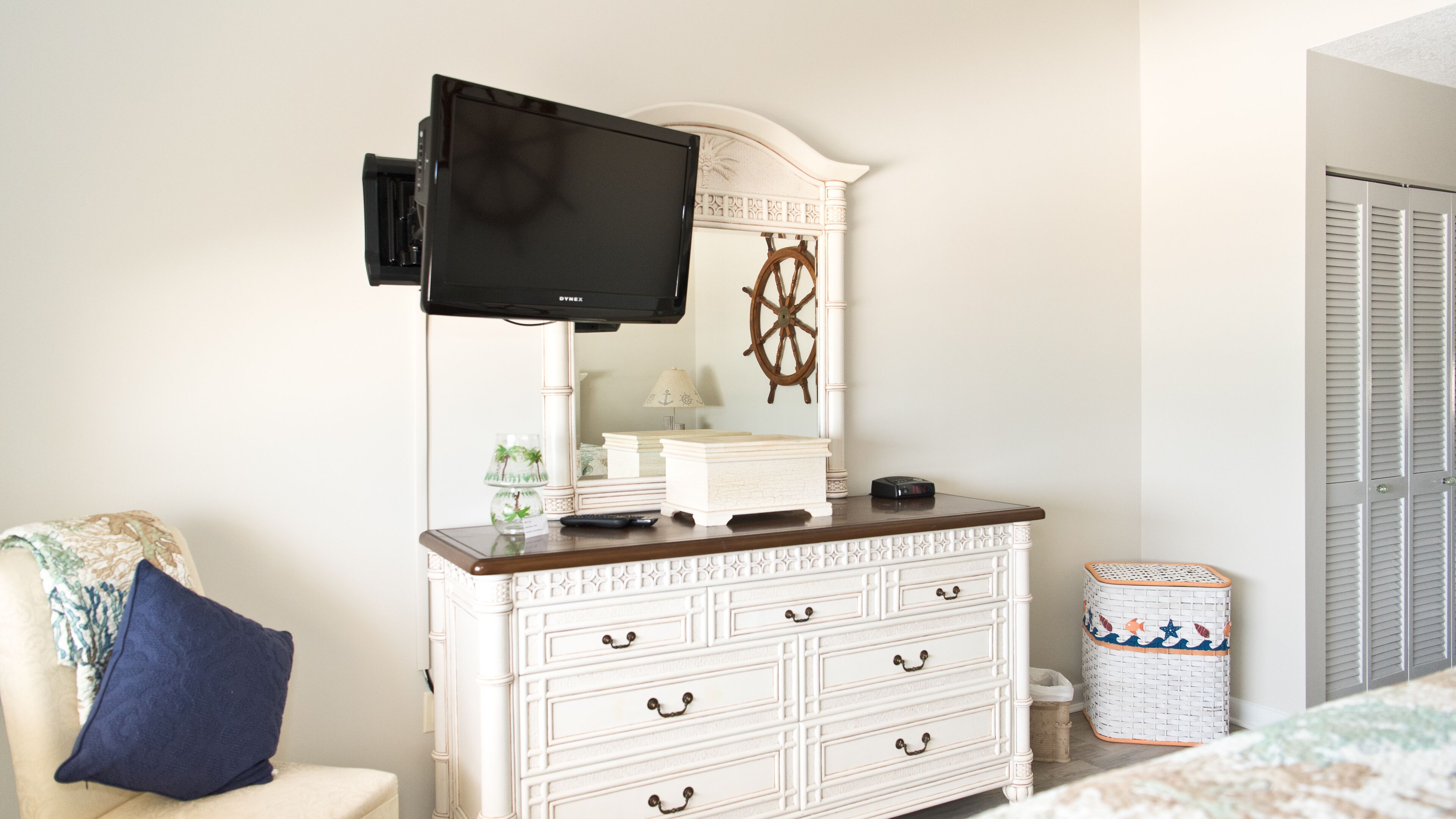 Une chambre avec une commode blanche, un téléviseur fixé au-dessus, une roue décorative et un lit recouvert d’une couvre-lit à motifs.