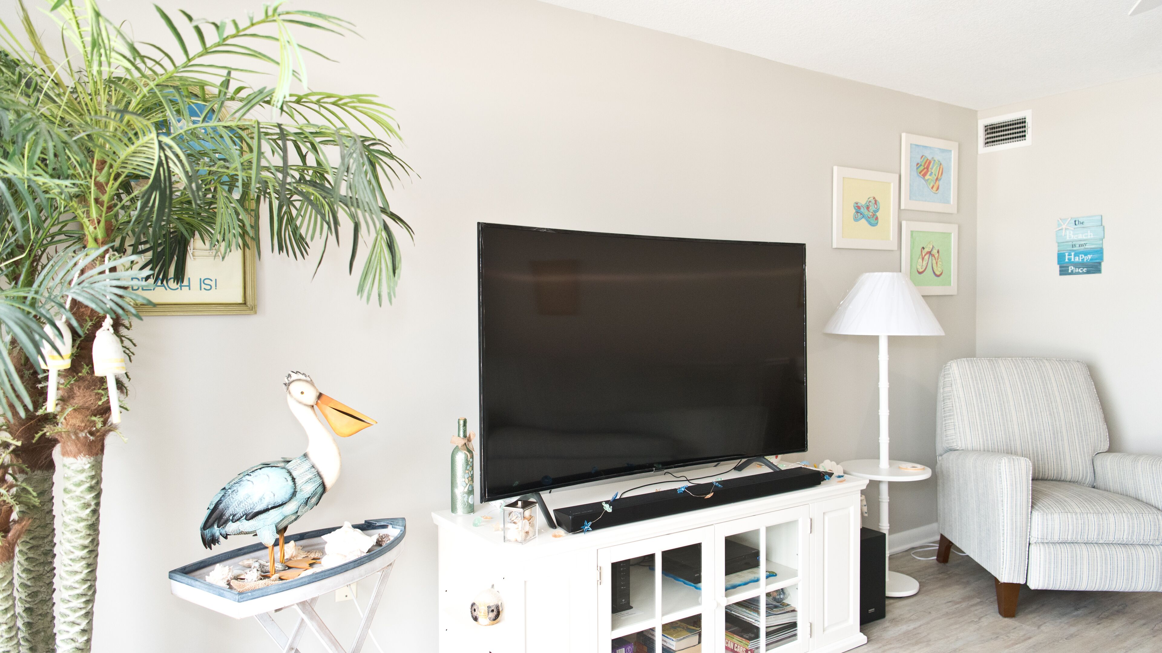 A living room with a flat-screen TV, a white cabinet, a striped armchair, a lamp, and a decorative bird statue.