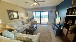 A living room with a sofa, coffee table, and a bookshelf. There is a view of the ocean through the sliding glass door. - Oceanfront 2 Bedroom Townhouse Summerhouse 305 (ST. AUGUSTINE)