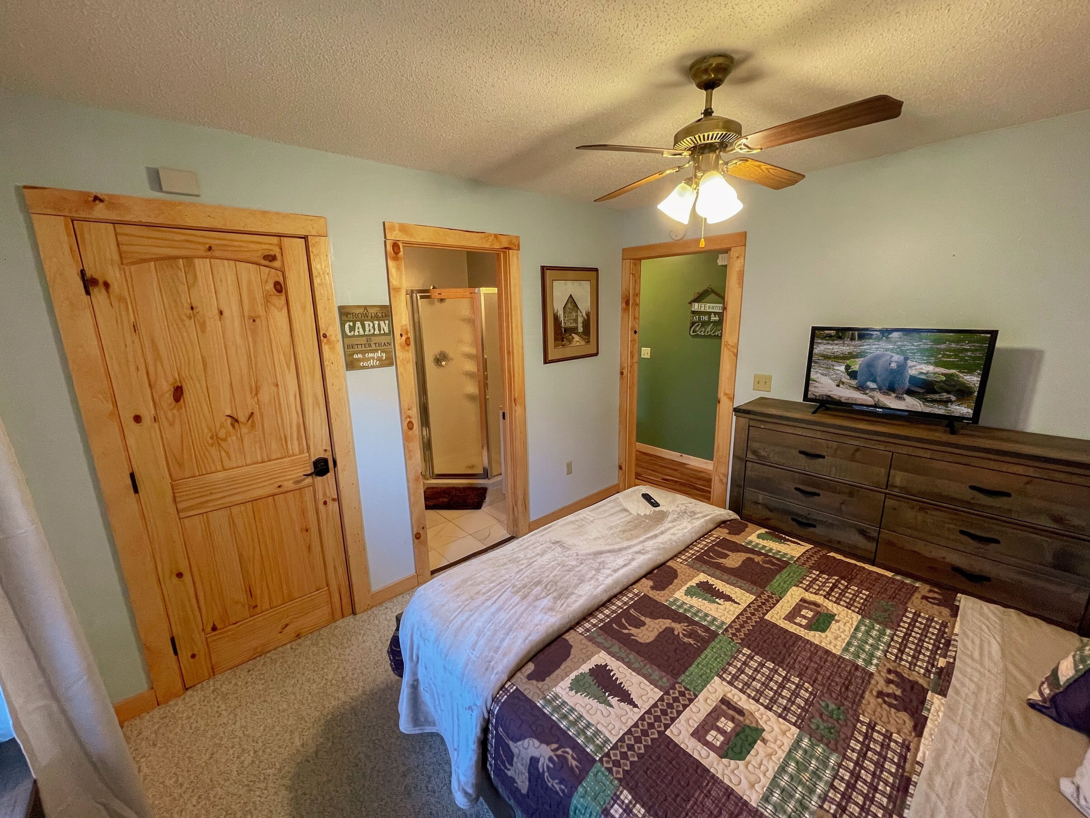 A bedroom with a wooden door, a bed with a quilt, a ceiling fan, a dresser, and a television on a stand.