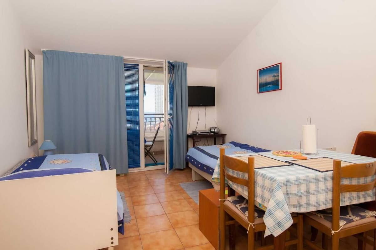 A small, compact room with two beds, a dining table, a TV, and a window with curtains.