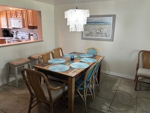 A dining area with a wooden table, blue placemats, wicker chairs, a hanging light fixture, and a beach-themed painting on the wall.