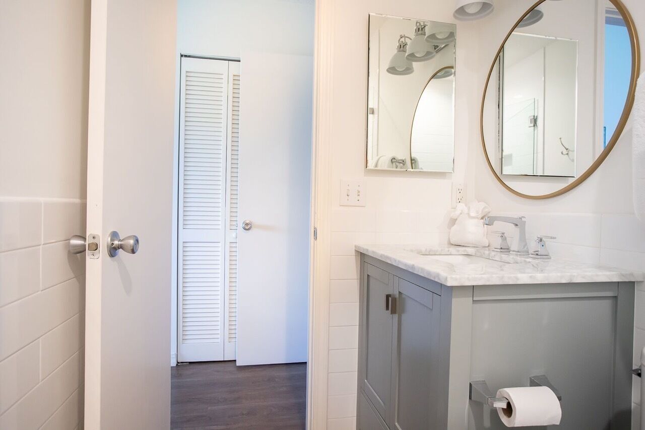 A bathroom with a white tiled wall, a gray vanity with a mirror, and a toilet paper holder.