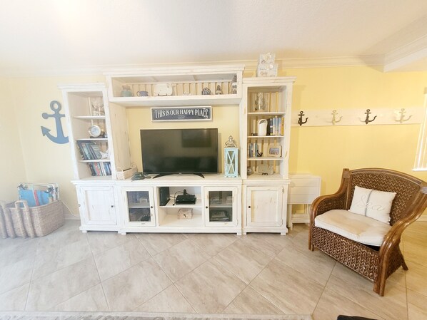 Sala de estar com televisão sobre um móvel branco, uma cadeira de vime e uma estante cheia de livros e objetos decorativos.