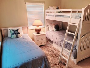 Une chambre avec deux lits simples, un lit superposé, une table de chevet avec une lampe et une fenêtre avec des stores.