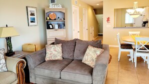 A living room with a grey sofa, a bookshelf, a dining table with chairs, and a kitchen area in the background. - Oceanfront 2 Bedroom Townhouse.  Summerhouse Unit 353 (ST AUGUSTINE)