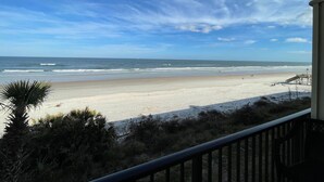 A beach view from a balcony with a railing, ocean, and sparse vegetation.