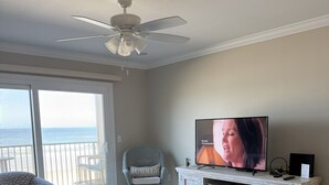 A living room with a television, a ceiling fan, a wicker chair, and a view of the ocean through a sliding glass door. - Oceanfront 2 Bedroom Townhouse.  Summerhouse Unit 357 (ST. AUGUSTINE)
