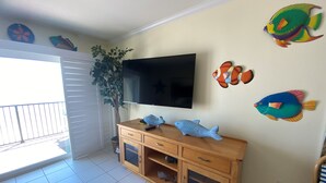 A living room with a flat-screen TV mounted on the wall, a wooden cabinet with drawers, a potted plant, and colorful fish wall decals.