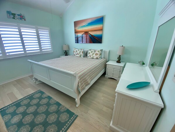 Un dormitorio con cama, mesitas de noche, cómoda, espejo, un cuadro y ventana con persianas.