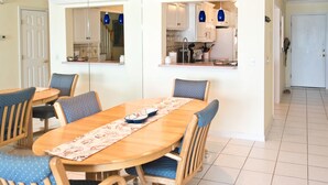 A dining area with a wooden table and blue upholstered chairs, a kitchen with white cabinets and blue pendant lights, and a tiled floor.