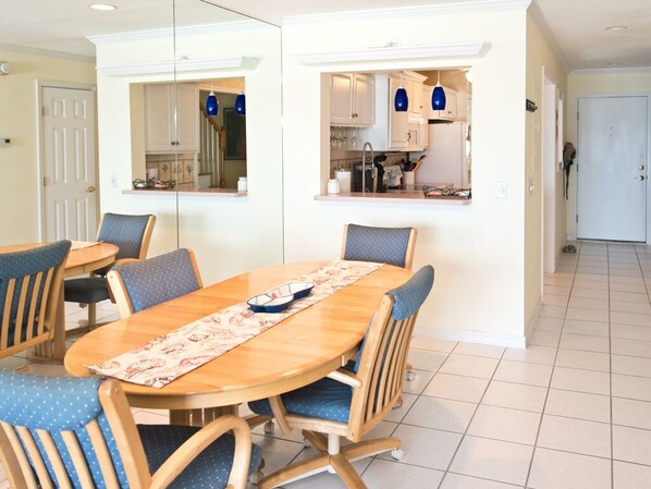 Un comedor con una mesa de madera y sillas tapizadas en azul, una cocina con armarios blancos y lámparas colgantes azules, y un suelo de baldosas.