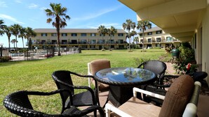 Un espace extérieur aménagé avec des chaises en osier et une table en verre, offrant une vue sur une piscine et un immeuble doté de nombreux balcons.