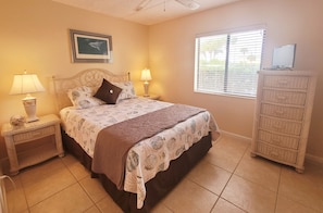 A bedroom with a bed, two bedside tables with lamps, a dresser, a window with blinds, and a framed picture on the wall.