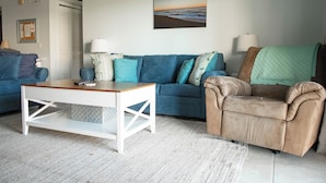 A living room with a blue sofa, a brown armchair, a white coffee table, and a painting of a beach scene.