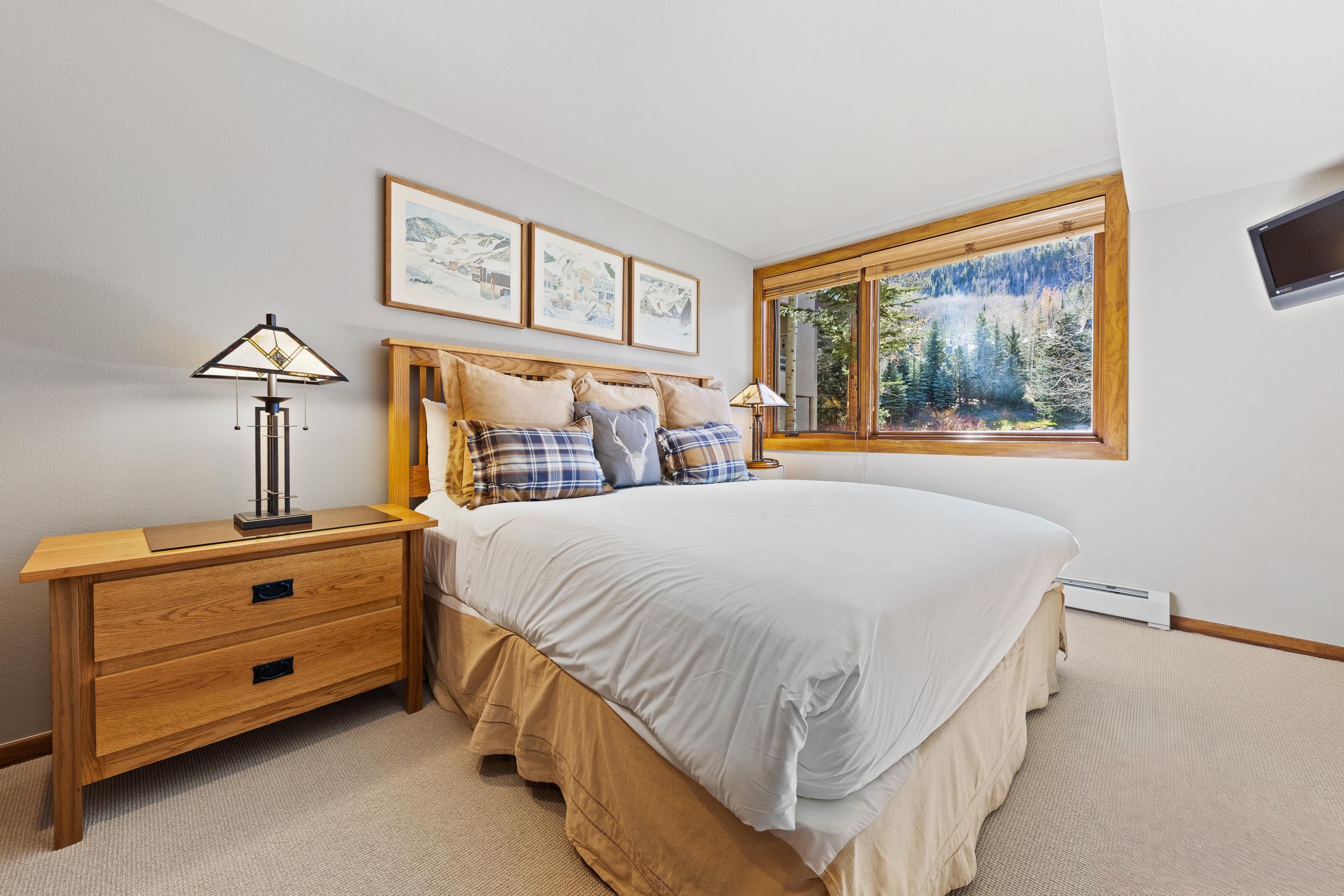 Ein Schlafzimmer mit einem großen Bett, einem hölzernen Nachttisch, einem Fenster mit Blick auf einen Wald und drei gerahmten Bildern an der Wand.