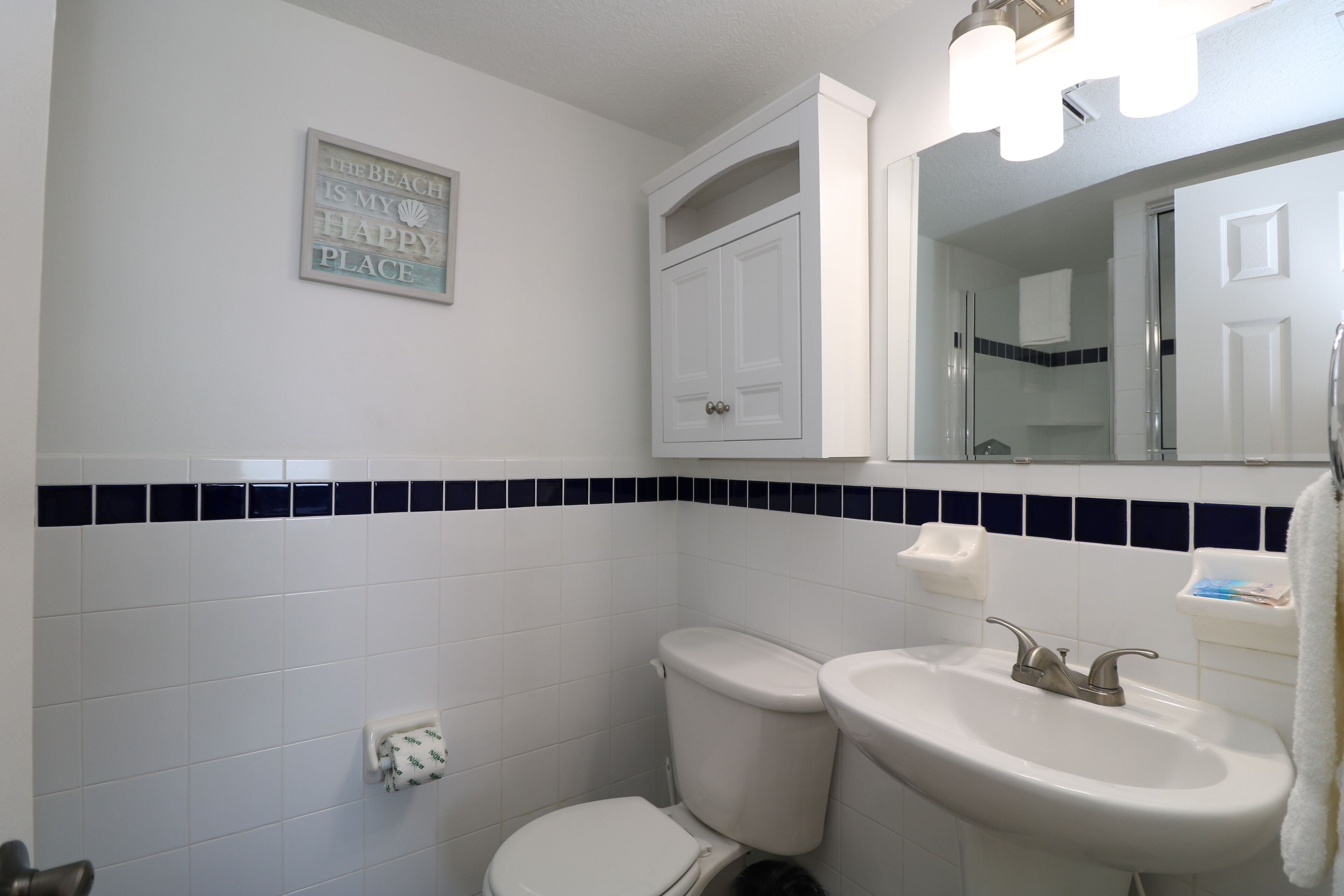 A bathroom with a toilet, sink, and a mirror above the sink. There is a towel rack and a sign on the wall.
