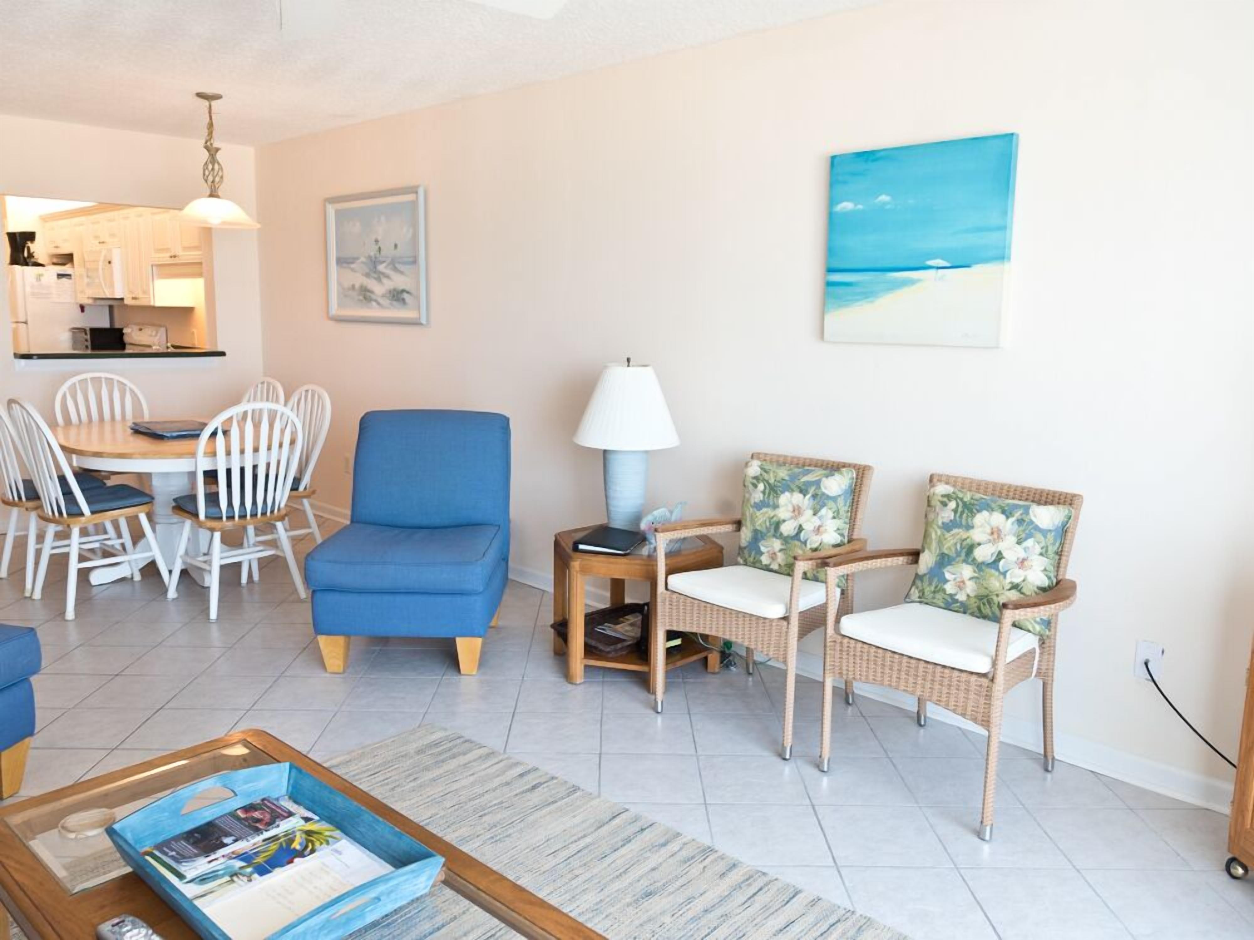 A living room with a dining area, a blue armchair, wicker chairs, a coffee table, and a painting of a beach.