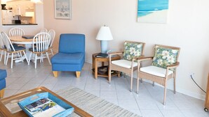 A living room with a dining area, a blue armchair, wicker chairs, a coffee table, and a painting of a beach. - Oceanfront 2 Bedroom Townhouse  Summerhouse Unit 456 (ST. AUGUSTINE)