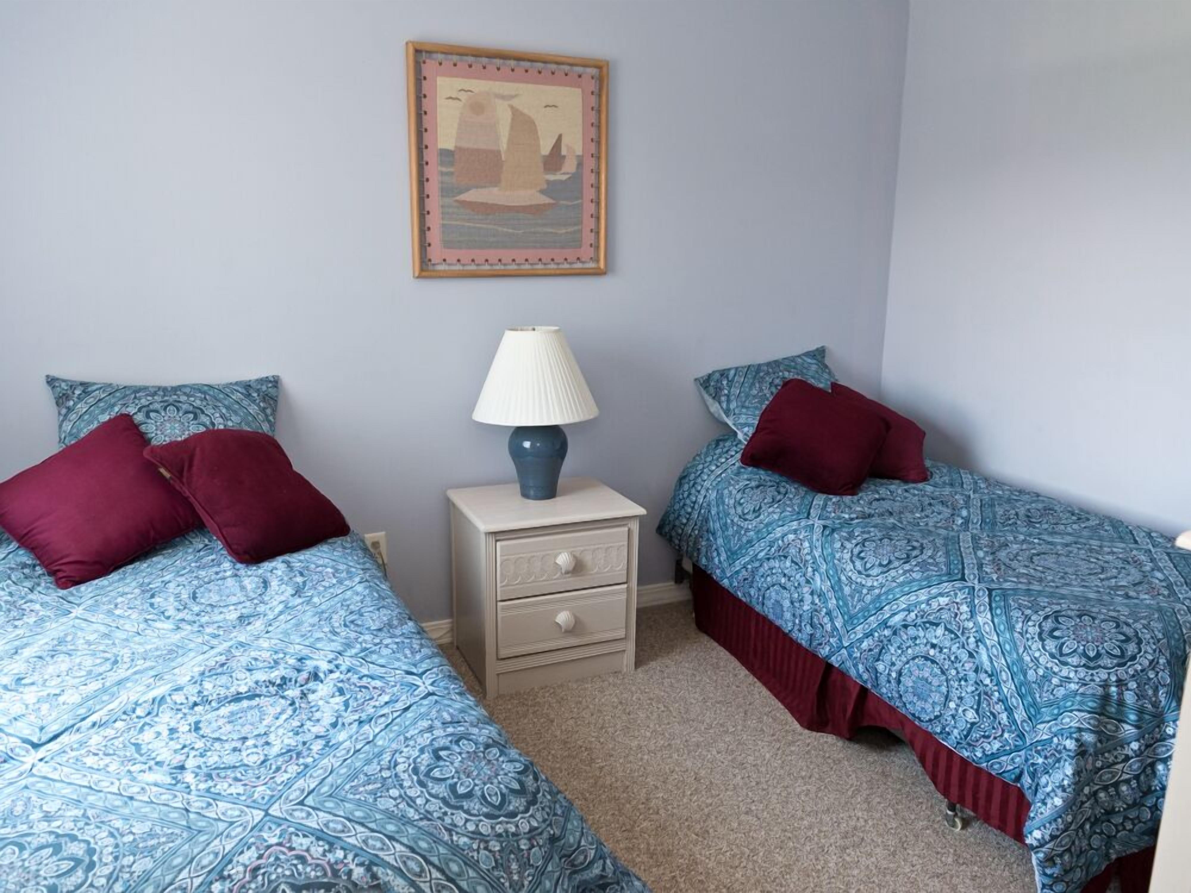 Une chambre avec deux lits, chacun recouvert d’une couvre-lit à motifs bleus et agrémenté d’oreillers rouges. Une table de chevet avec une lampe et un tiroir se trouve entre les deux lits. Un tableau encadré est accroché au mur, au-dessus des lits.