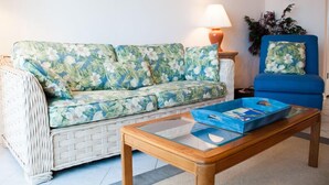 A living room with a floral sofa, a blue armchair, a wooden coffee table, a lamp, and a framed picture on the wall. - Oceanfront 2 Bedroom Townhouse  Summerhouse Unit 456 (ST. AUGUSTINE)