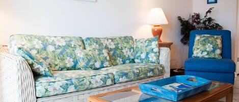 A living room with a floral sofa, a blue armchair, a wooden coffee table, a lamp, and a framed picture on the wall.