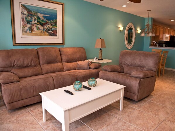 A living room with a brown leather sofa, a white coffee table, and a painting of a coastal scene.