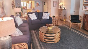 A cozy living room with a dark blue sofa, a glass coffee table, and a wicker chair. There is a lamp on the left and a framed picture on the wall.