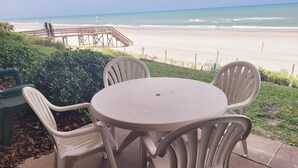 A patio with a round table and four chairs, overlooking a beach with a wooden structure in the background.