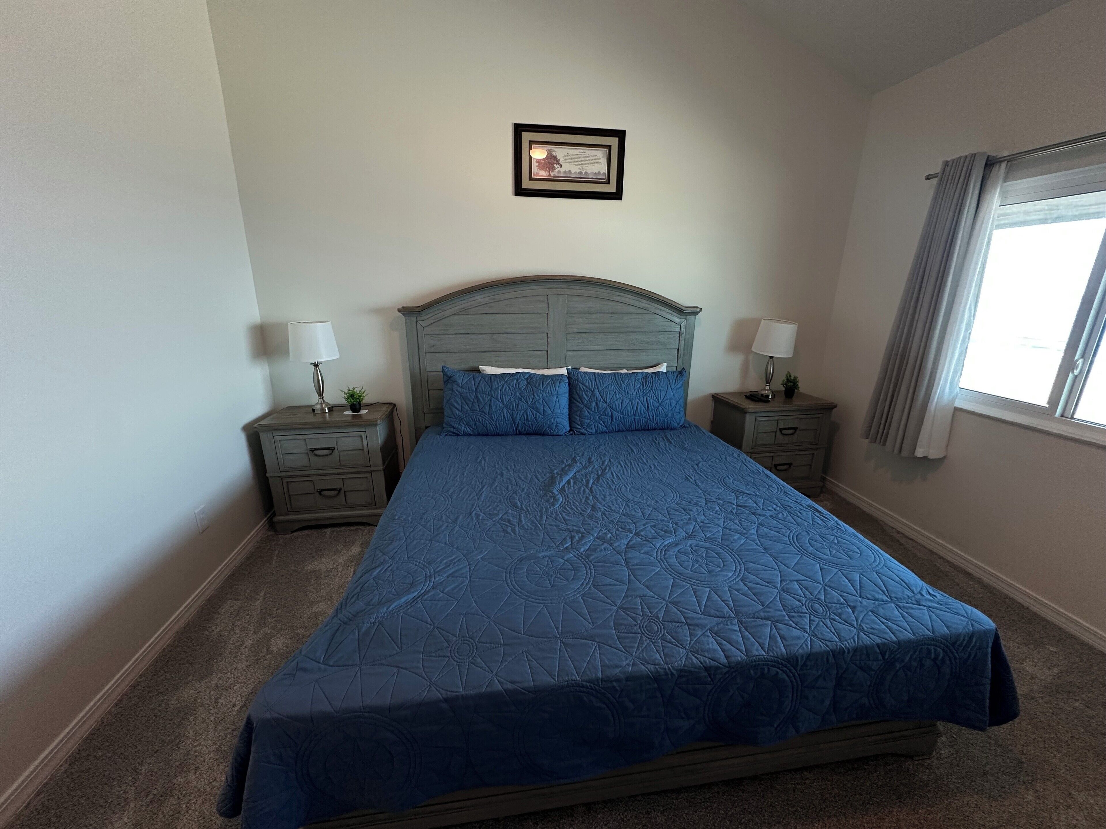 A bedroom with a large bed, two bedside tables, a nightstand, a framed certificate on the wall, and a window with curtains.