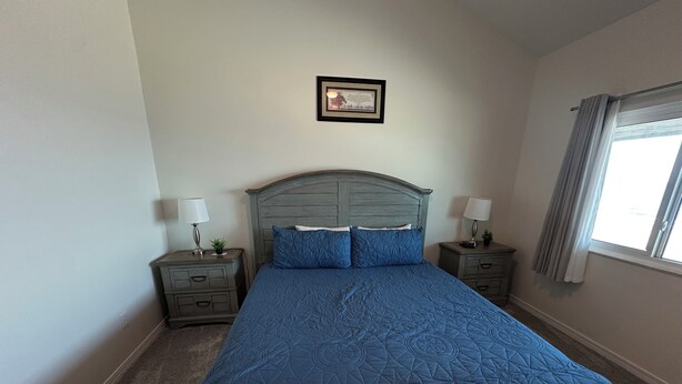 Une chambre avec un grand lit, deux tables de chevet, une commode, un certificat encadré accroché au mur et une fenêtre avec des rideaux.
