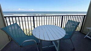 Un balcon avec une table et deux chaises, donnant sur une plage où l’on peut admirer les vagues et un ciel dégagé.