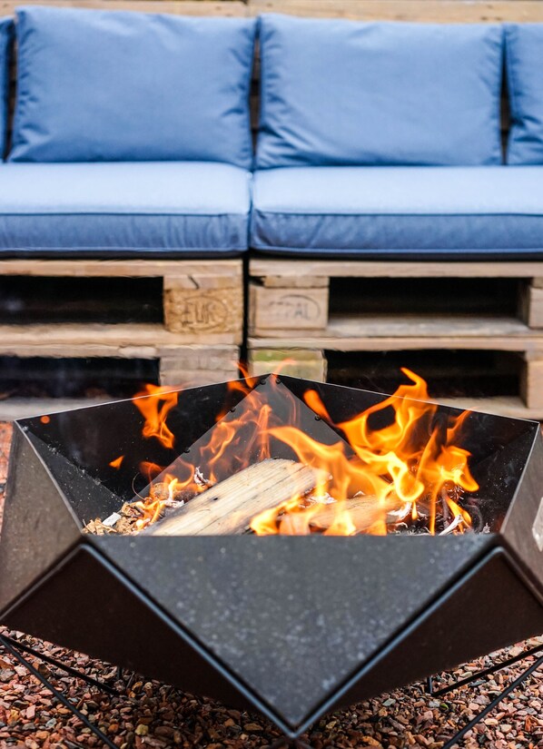 A fire pit with logs and flames, surrounded by a blue sofa and wooden crates.