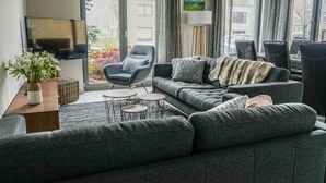 Ein modernes Wohnzimmer mit grauem Sofa, grauem Sessel und einem kleinen Couchtisch. An der Wand ist ein Fernseher angebracht, eine Stehlampe steht bereit und ein Fenster mit Vorhängen sorgt für Licht.