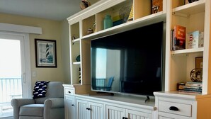 A living room with a large flat-screen TV, a white entertainment center, a patterned rug, a gray armchair, and a framed picture on the wall. - Beautiful Direct Oceanfront 2 bed 2.5 bath Townhouse- Summerhouse 162 (St. Augustine)