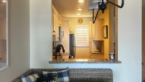 A cozy living area with a wooden bench, a wicker sofa, and a dining table. A kitchen is visible in the background with modern appliances and a large window. - Beautiful Direct Oceanfront 2 bed 2.5 bath Townhouse- Summerhouse 162 (St. Augustine)