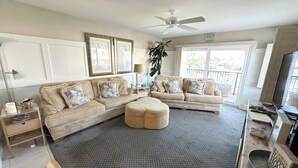 Un salon avec un ensemble de canapés de couleur beige, une table basse, une table d’appoint et un lampadaire. On y trouve également un ventilateur de plafond, une grande fenêtre donnant sur l’extérieur et un téléviseur posé sur un meuble.
