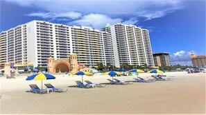 Um hotel à beira-mar com vários edifícios altos, uma entrada em arco destacada e diversos guarda-sóis e espreguiçadeiras de praia.