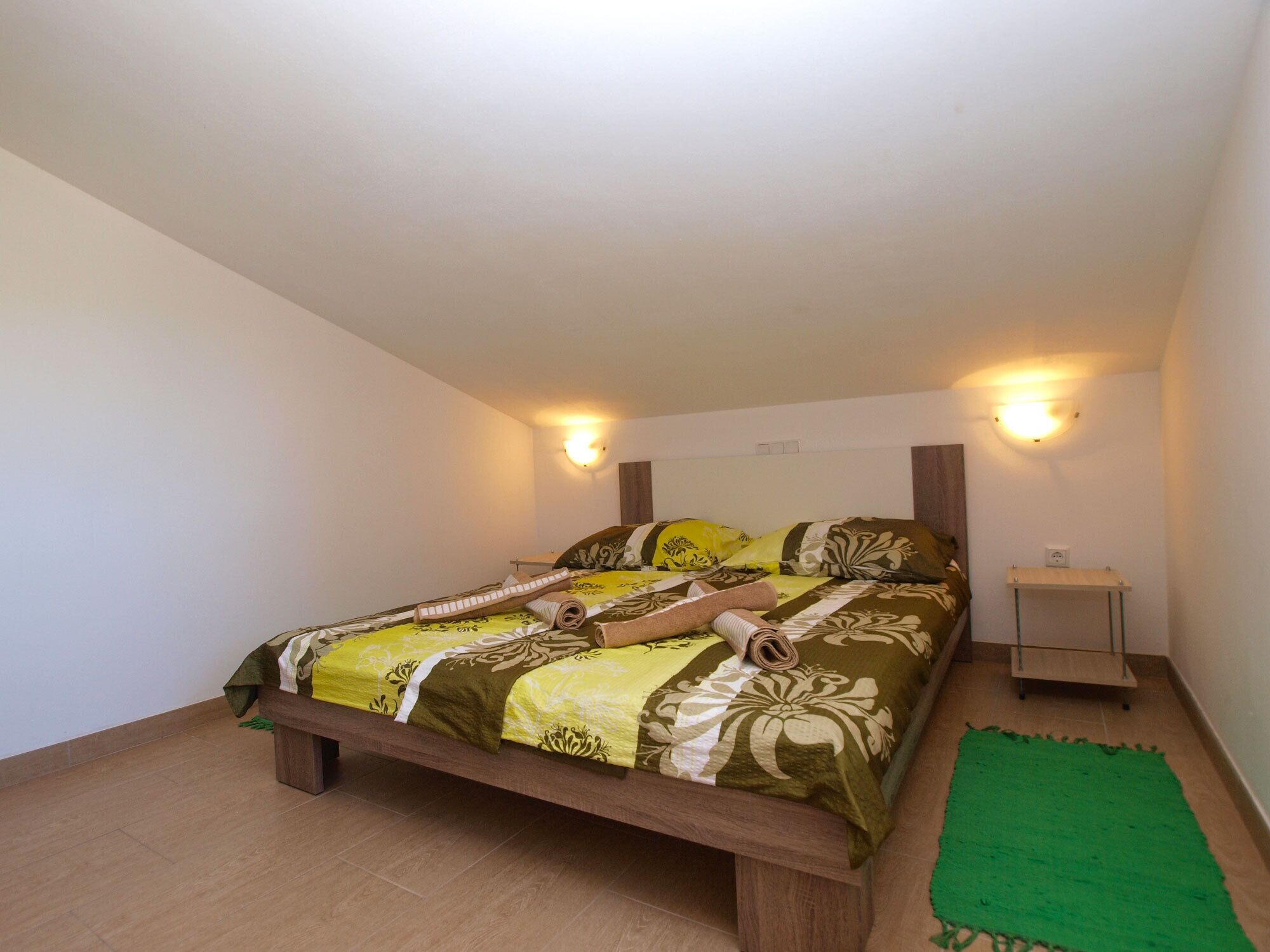 A single-bed room with a wooden frame, a green rug, and two bedside tables with wall-mounted lamps.