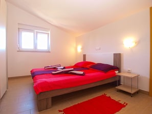 Un dormitorio con cama de madera, ropa de cama roja, una mesita de noche pequeña y una ventana con persianas.