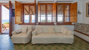 A room with a sofa, a bench, and a view through open wooden shutters. - Villa Timbora on the Beach with Ocean View, Terrace (Cabras)