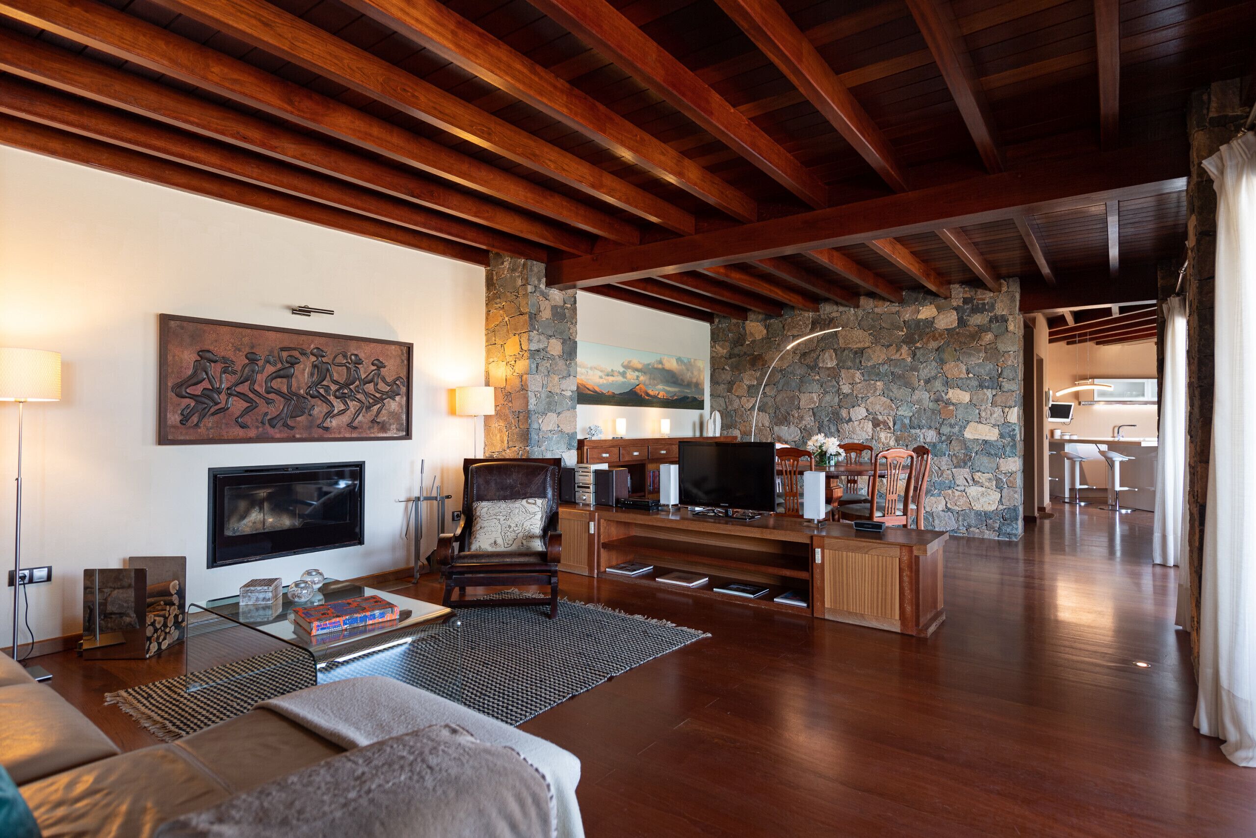 Amplia sala de estar con pared de piedra, vigas de madera en el techo, chimenea, televisor y comedor con sillas.