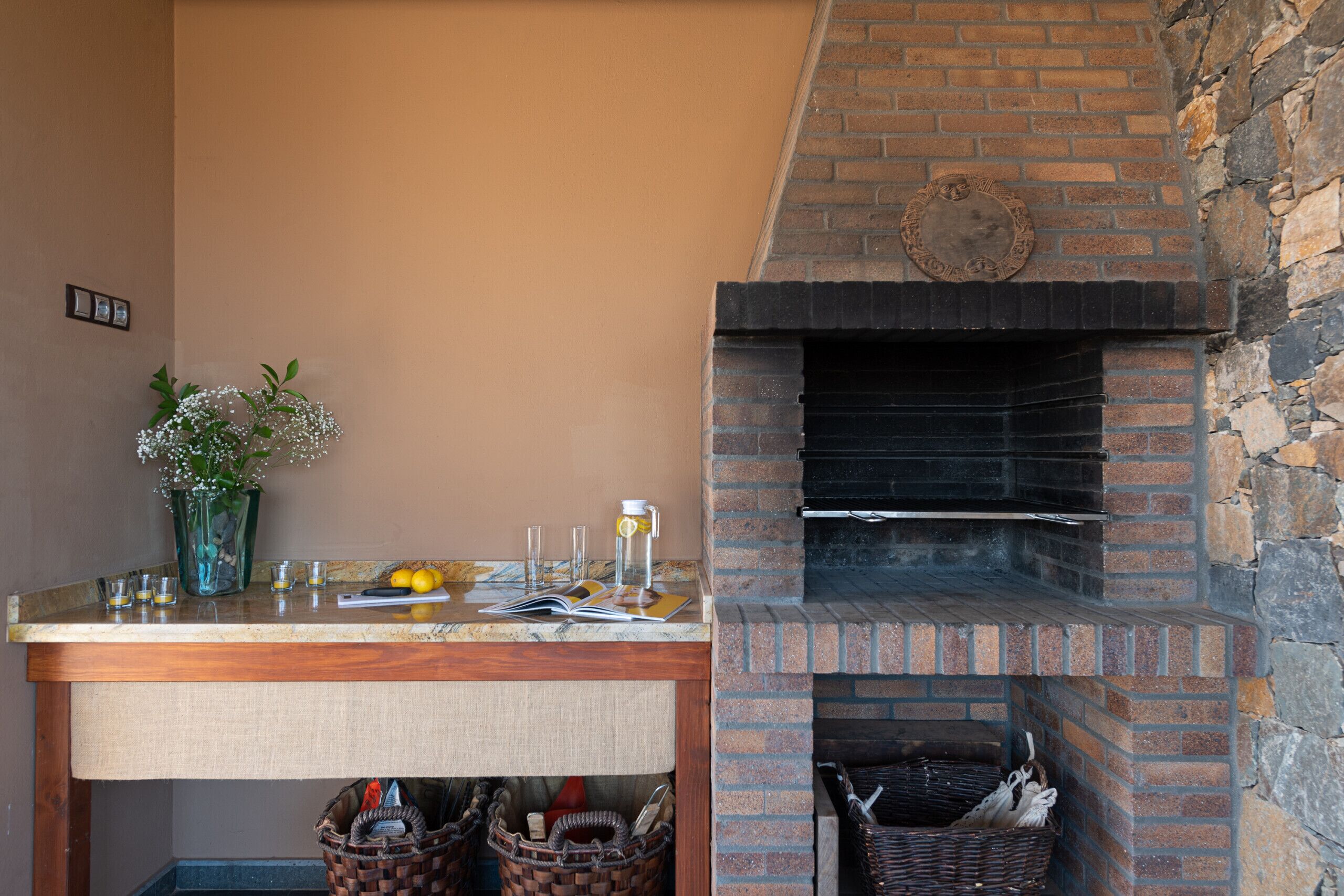 Una chimenea de ladrillo con pared de piedra, una mesa de madera con un florero y cestas debajo.
