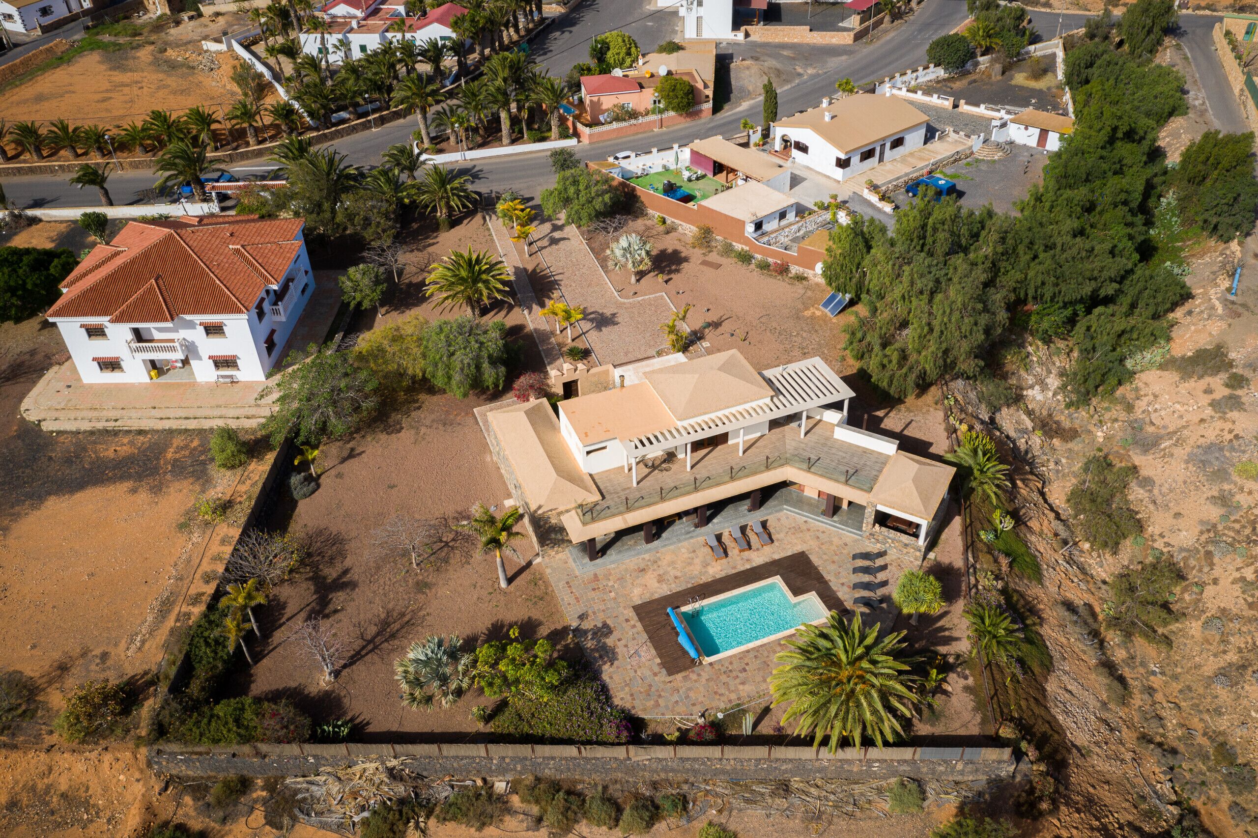 Vue aérienne d’un quartier résidentiel, avec une maison dotée d’une piscine, entourée de palmiers et d’un paysage vallonné.