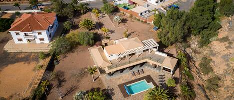 Vue aérienne d’un quartier résidentiel, avec une maison dotée d’une piscine, entourée de palmiers et d’un paysage vallonné.