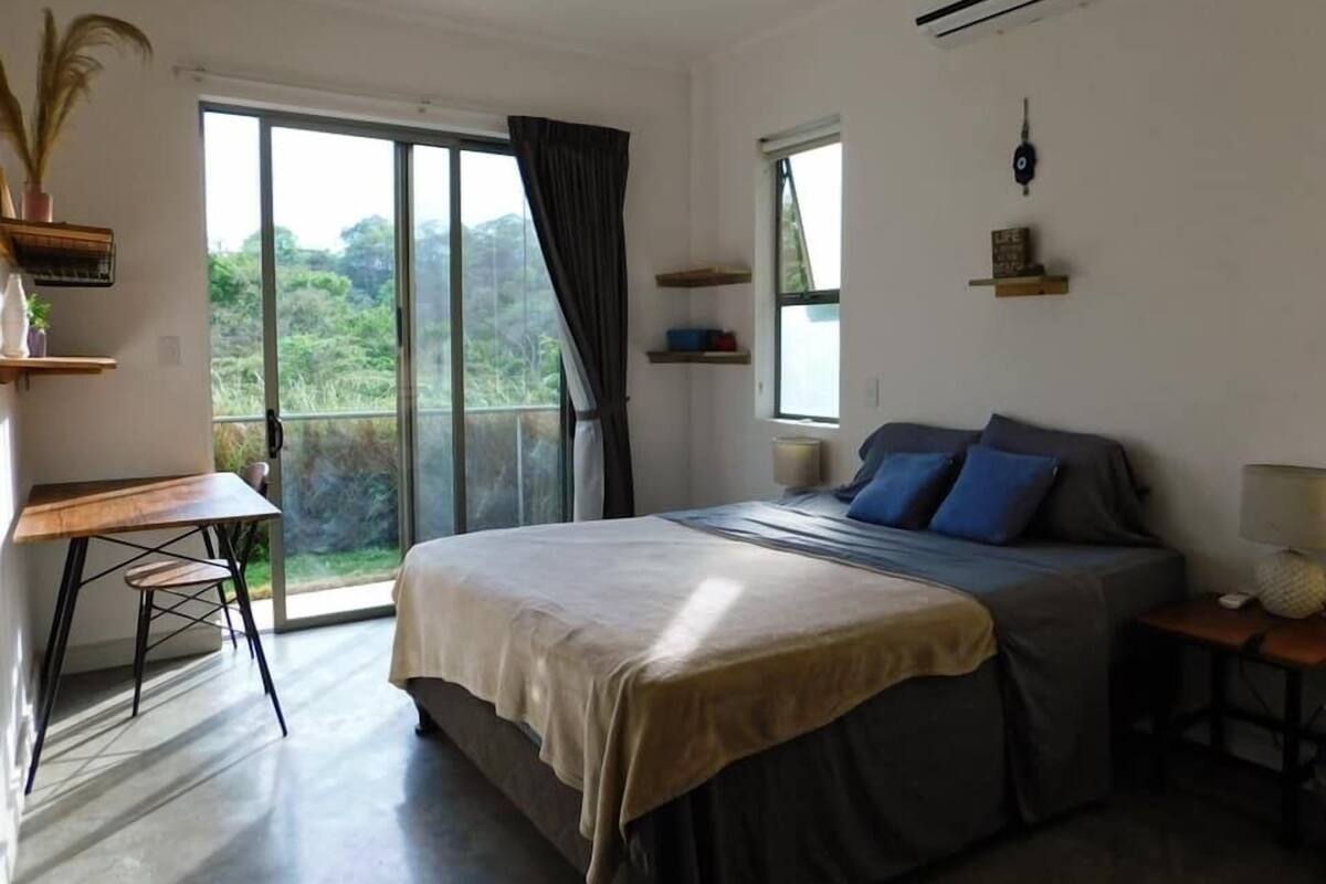 Quarto com cama, escrivaninha e cadeira. Janela com vista para o verde lá fora.